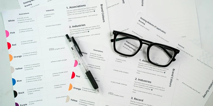 A desk covered with printed branding survey forms, showing color choices, industry categories, and brand associations. A black pen and a pair of black-framed glasses rest on top of the papers.
