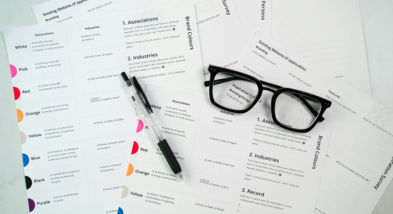 A desk covered with printed branding survey forms, showing color choices, industry categories, and brand associations. A black pen and a pair of black-framed glasses rest on top of the papers.