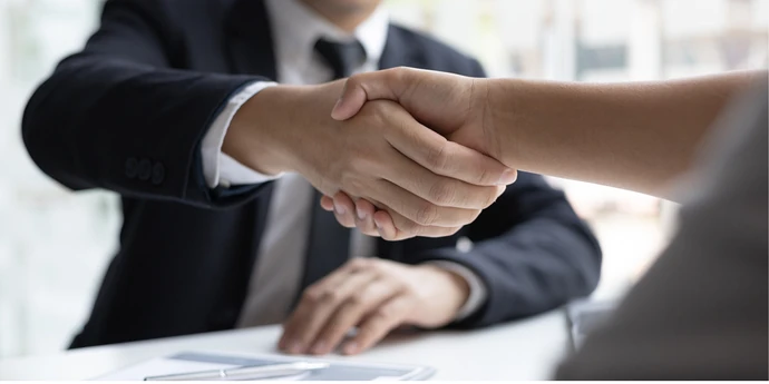 Two people shaking hands across a desk in an office.