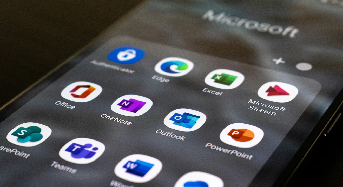 A close-up of a smartphone displaying a folder of Microsoft apps, including Word, Excel, Outlook, PowerPoint, Teams, OneNote, and OneDrive.