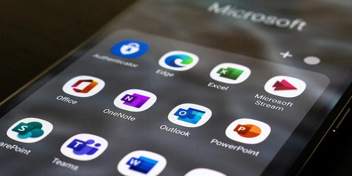 A close-up of a smartphone displaying a folder of Microsoft apps, including Word, Excel, Outlook, PowerPoint, Teams, OneNote, and OneDrive.
