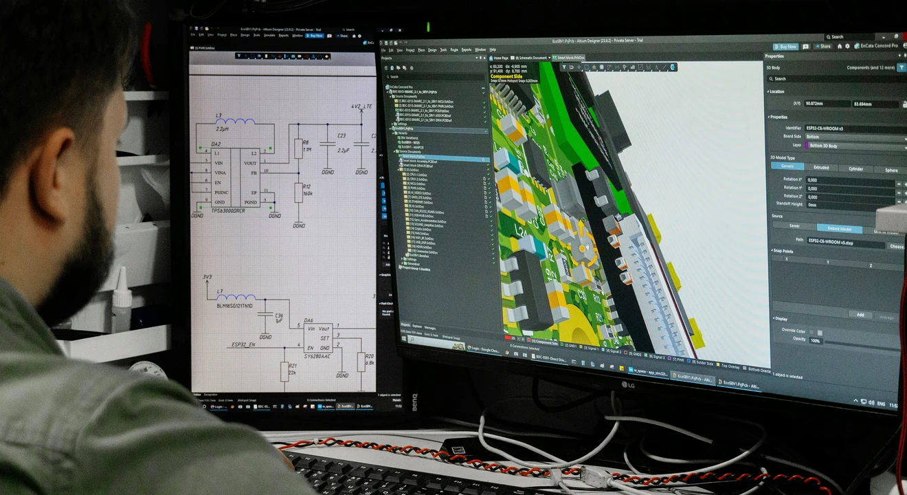 Person working at a dual-monitor setup, viewing circuit diagrams on one screen and a 3D PCB design on the other, in a technical workspace