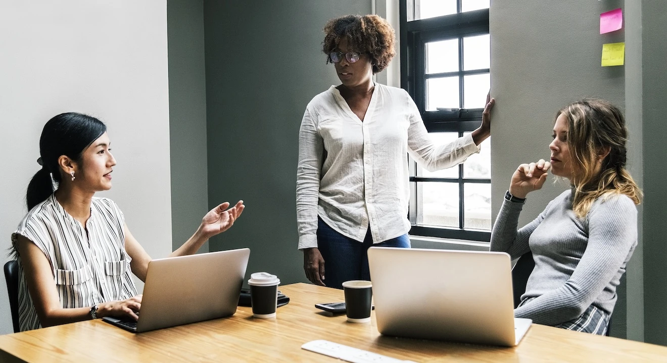 Three women collaborating in a meeting room with laptops, discussing work around a table