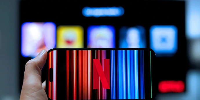 A hand holds a smartphone displaying the Netflix logo against a colorful striped background, with a blurred streaming interface visible on a screen behind it.