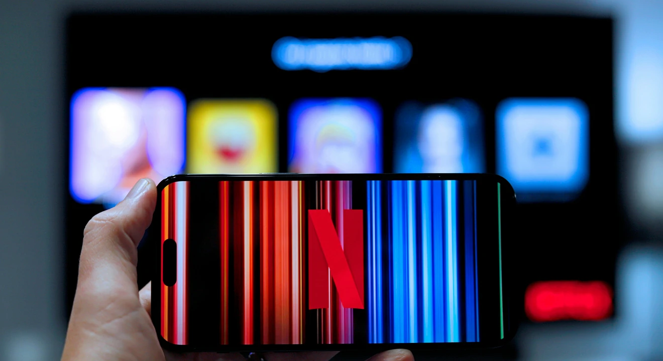 A hand holds a smartphone displaying the Netflix logo against a colorful striped background, with a blurred streaming interface visible on a screen behind it.