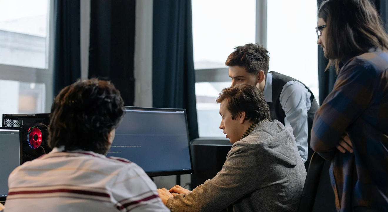 A group of people gather around a man sitting in front of several computers in an office conference room