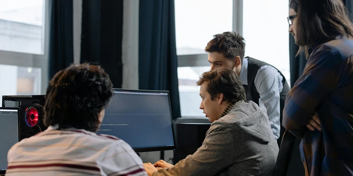A group of people gather around a man sitting in front of several computers in an office conference room