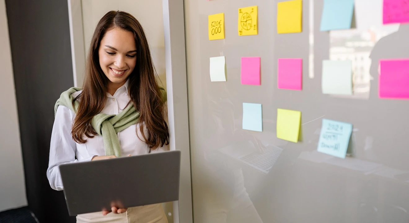 Woman using a laptop beside a glass wall covered with colorful sticky notes