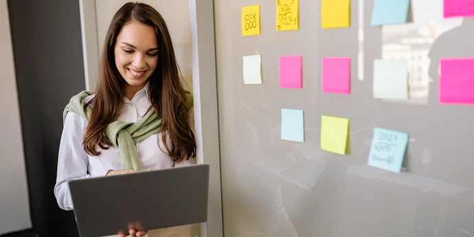 Woman using a laptop beside a glass wall covered with colorful sticky notes