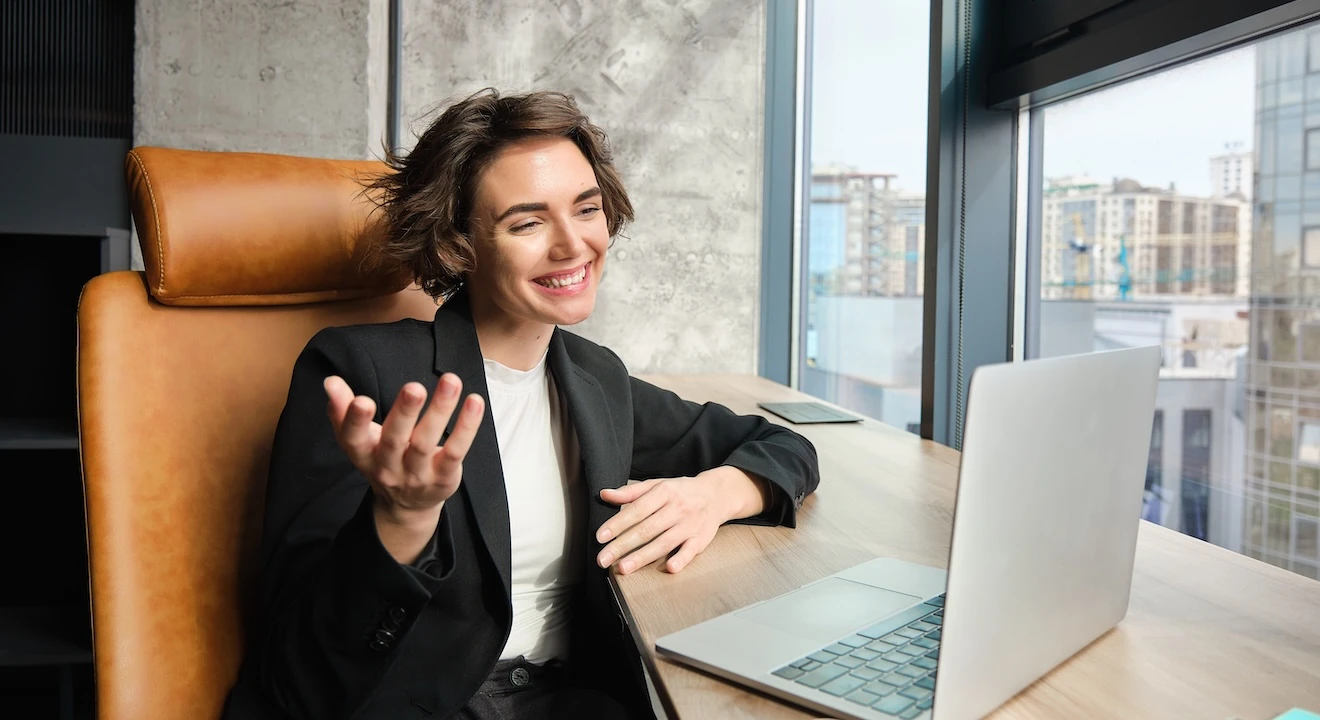Professional woman in a blazer speaking and gesturing during a video call on a laptop in an office setting