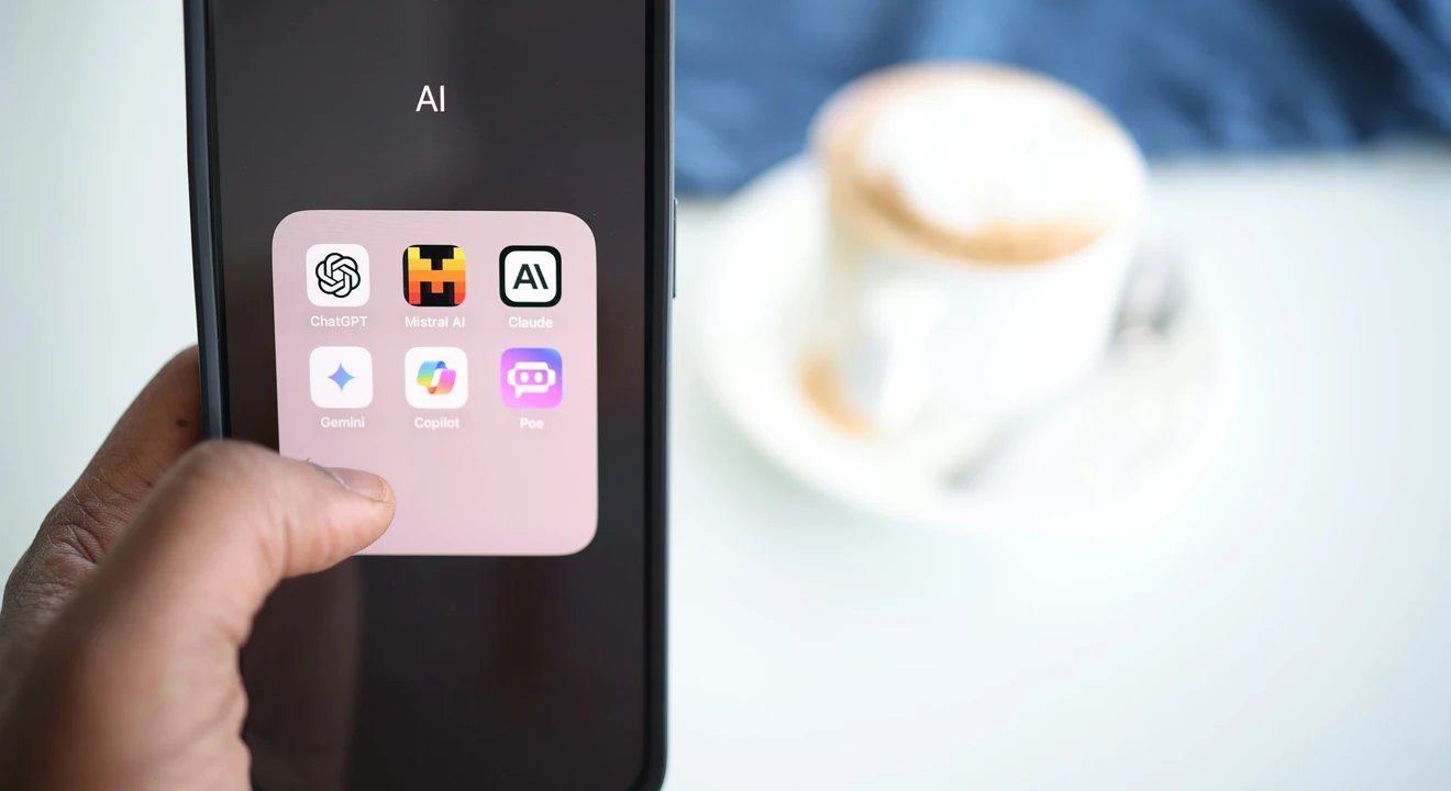 Smartphone screen showing a folder of AI apps including ChatGPT, Claude, Gemini, Copilot, and Poe, held in hand with a blurred coffee cup in the background.