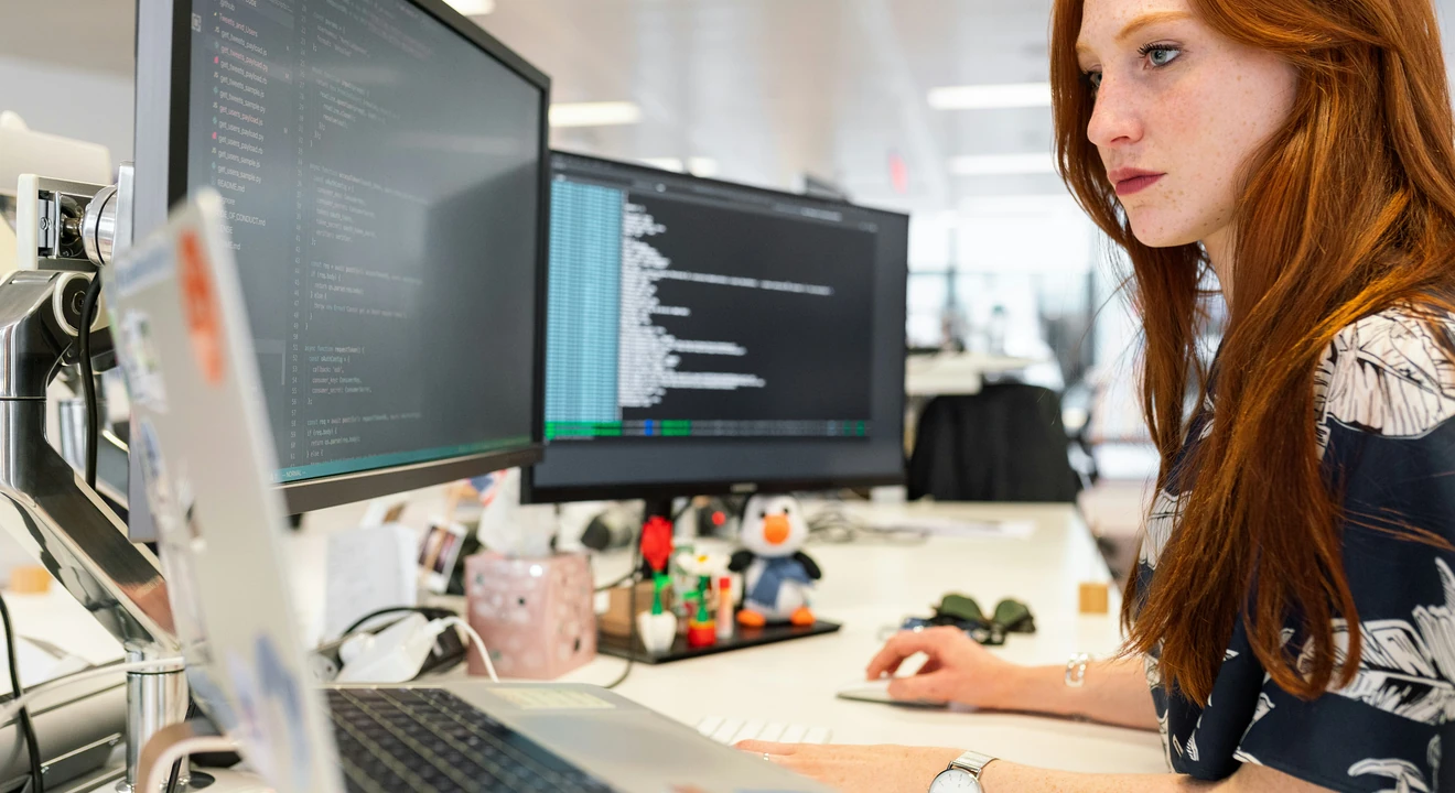 Person working at a desk with dual monitors displaying code, using a laptop in an office environment